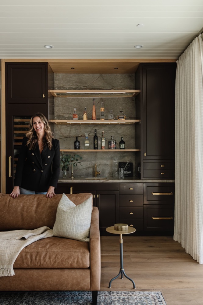 Daniela Z in a home bar she designed, with dark cabinetry, stone backsplash and oak shelving